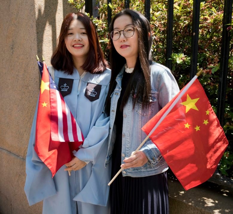 Two young Chinese women with dark hair hold red flags with yellow stars on them. One of the women wears a light blue graduation robe and smiles.